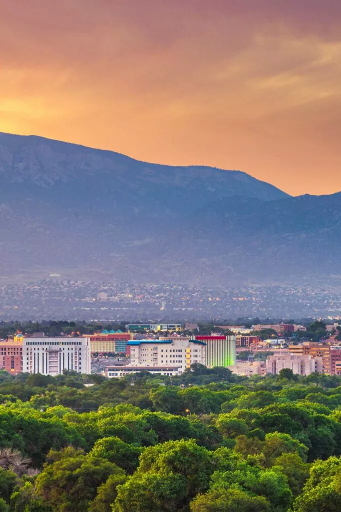 Albuquerque skyline glowing at sunset with the Sandia Mountains behind and green cottonwood bosque in the foreground