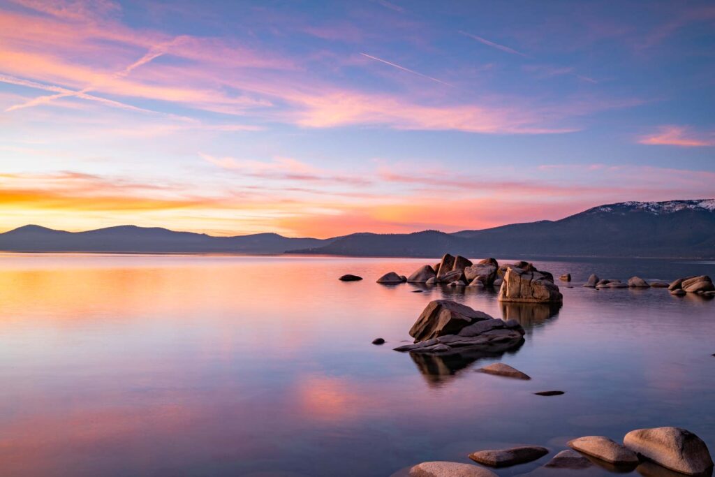Lake Tahoe at sunset with pink and orange sky reflected on calm water and granite boulders, Sierra Nevada peaks in silhouette, about 45 minutes from Reno