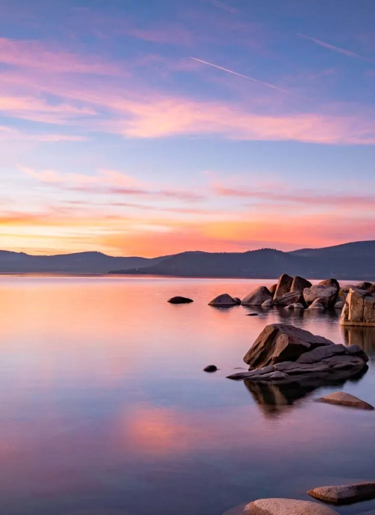 Lake Tahoe at sunset with pink and orange sky reflected on calm water and granite boulders, Sierra Nevada peaks in silhouette, about 45 minutes from Reno